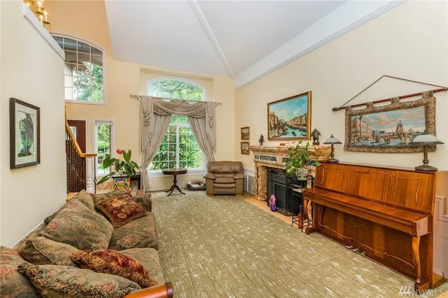The living room at A Rose Garden Home 1, with a vaulted ceiling, a piano, a marble fireplace, and a tall arched window onto greenery